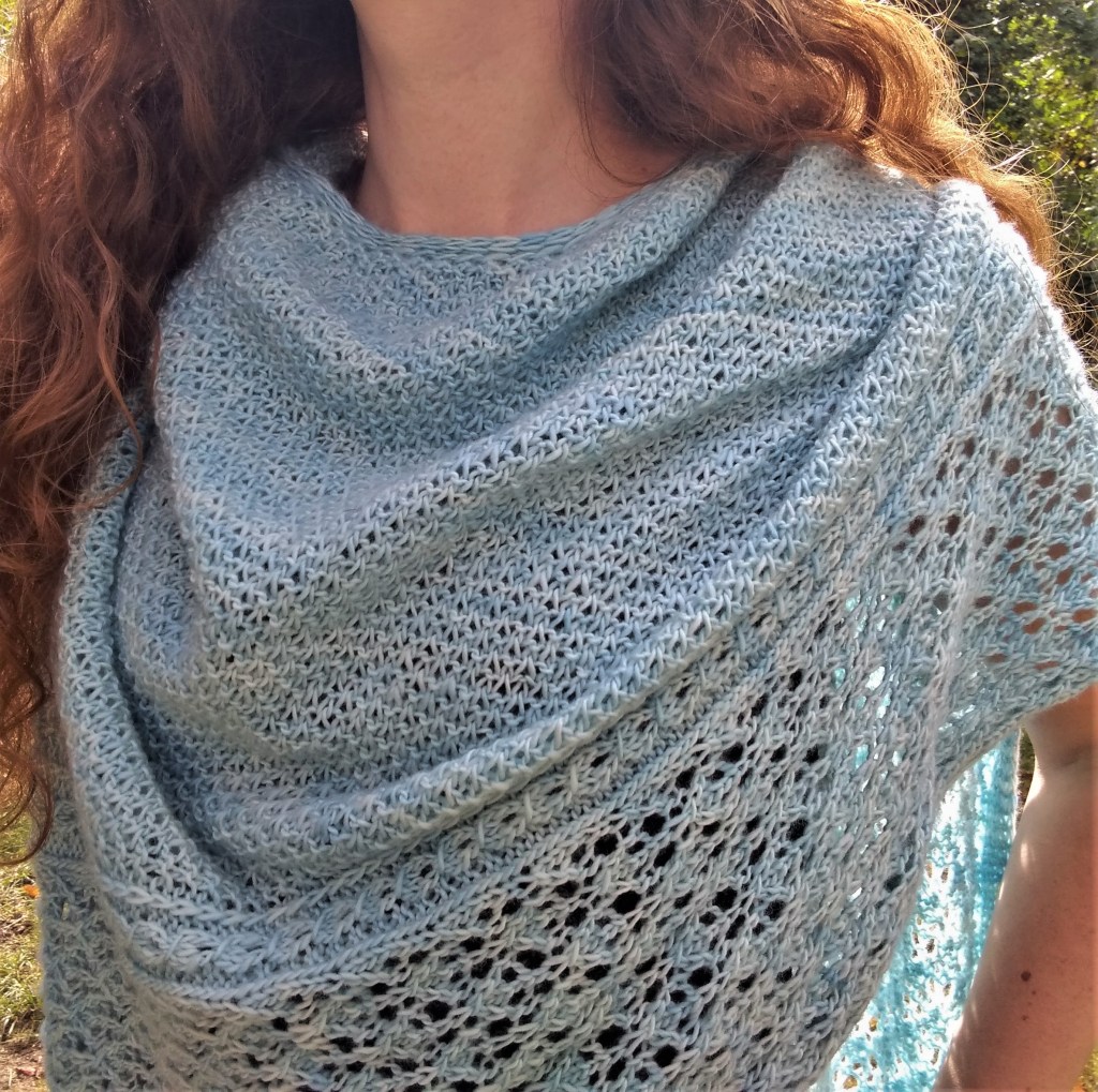 A richly textured, icy blue shawl with a lace border is draped around a person's neck. The person is standing outdoors on a sunny day.