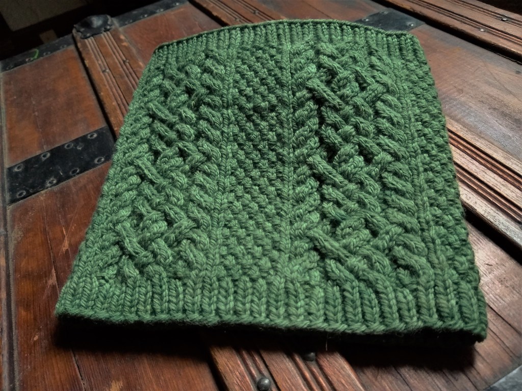 A green cowl with tightly woven cables surrounded by squishy textures is laying on an old wooden trunk.