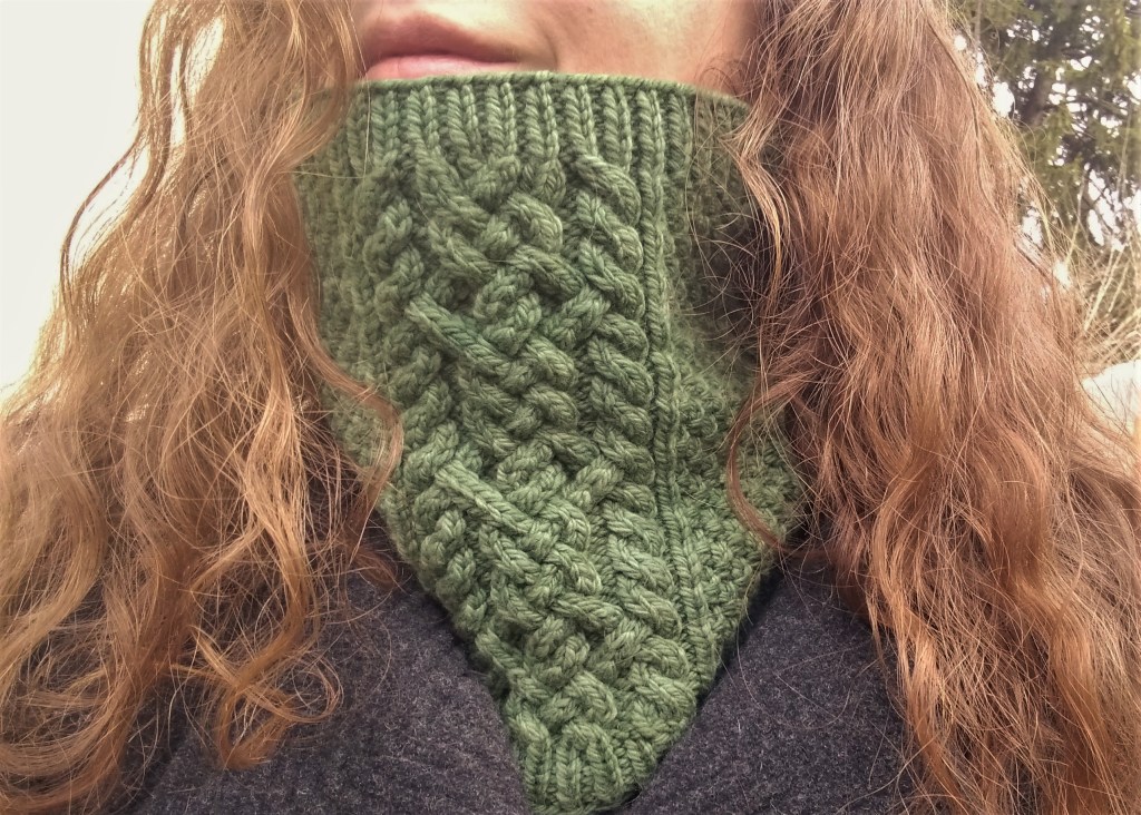 A woman with long, curly auburn hair wears a green cabled cowl around her neck.