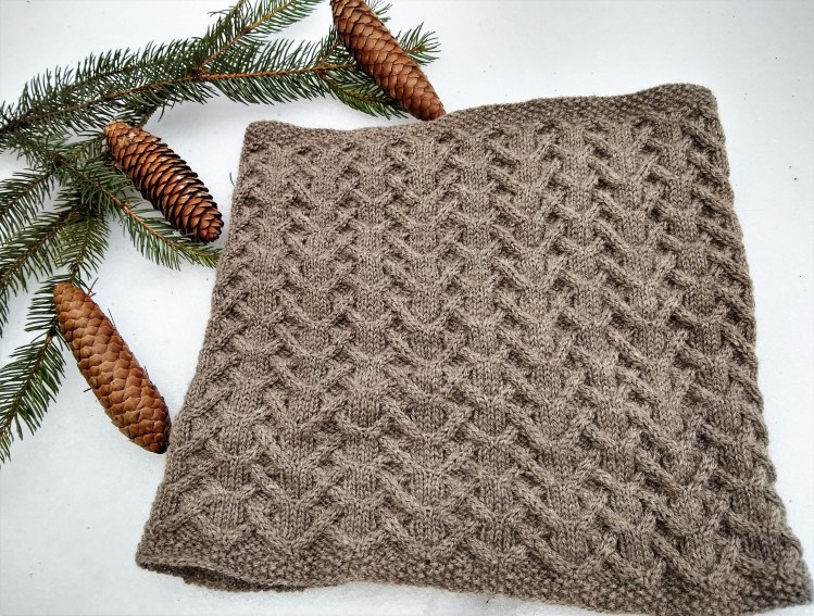 A densely cabled cowl knit in a warm gray colored yarn lays on top of fresh snow, with several pine boughs and cones by its upper left corner.